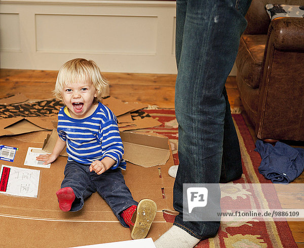 Junge sitzt lachend auf Pappkarton im Wohnzimmer