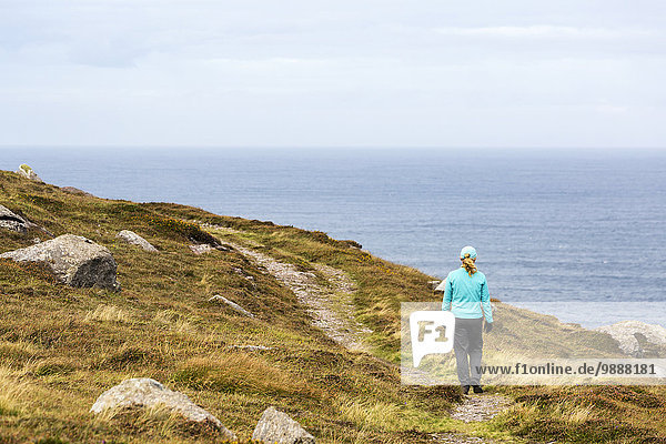 Female hiker along pathway of grassy hillside with rocks overlooking ocean; Ballinglanna  County Cork  Ireland