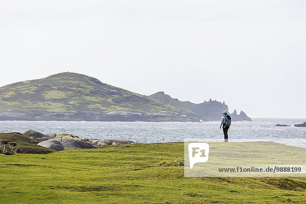 Female hiker on grassy field overlooking the ocean with large island in background; Omey Island  County Galway  Ireland
