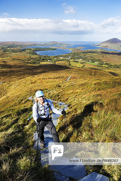 Female hiker climbing trail up grassy mountain with bay in the distance; Letterfrack  County Galway  Ireland