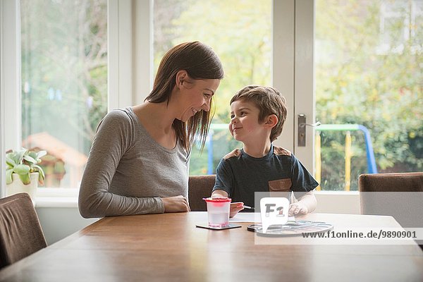 Boy smiling at mother as he paints on paper at table in living room
