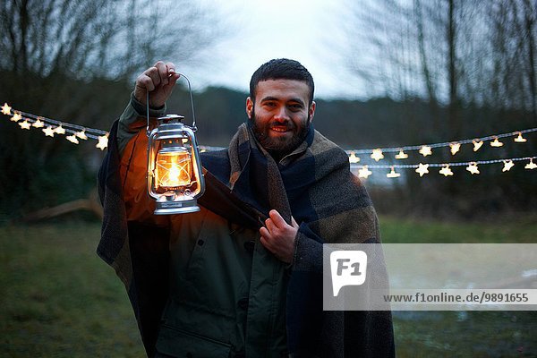 Young male camper holding up lantern whilst wrapped in blanket in woods