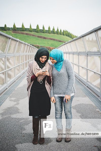 Two young female friends reading smartphone texts on park footbridge