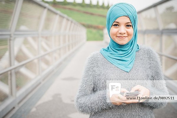 Porträt einer jungen Frau in türkisfarbenem Hijab mit Smartphone auf Steg
