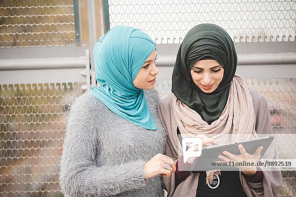 Two young women wearing hijabs using touchscreen on digital tablet on footbridge
