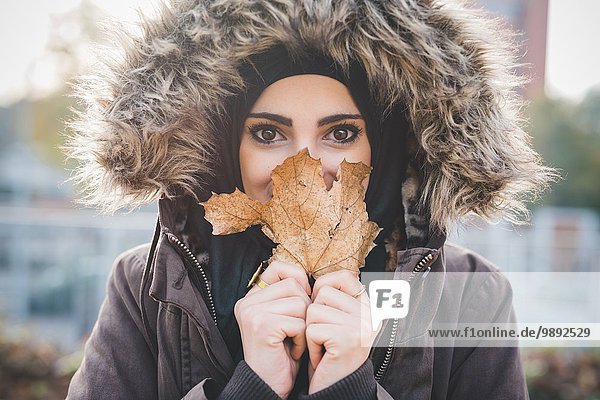 Close up of beautiful young woman in park holding up autumn leaf in front of face