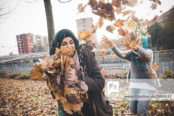 Two young women friends play fighting with autumn leaves in park