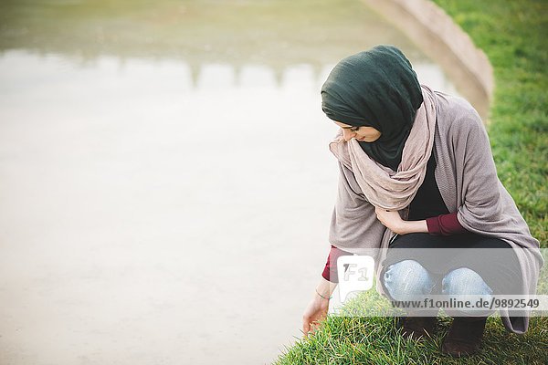 Young woman wearing hijab crouching next to park lake