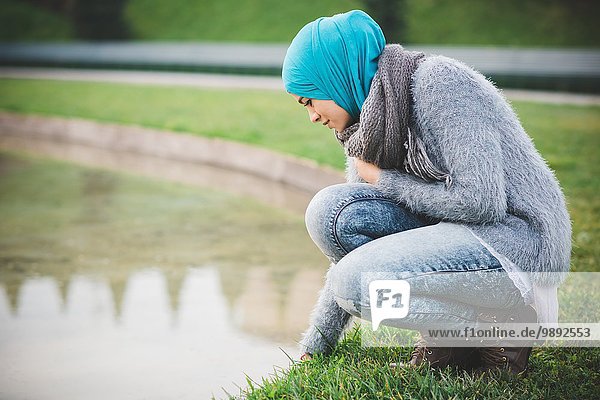 Junge Frau mit Hijab  die am Seeufer des Parks hockt.