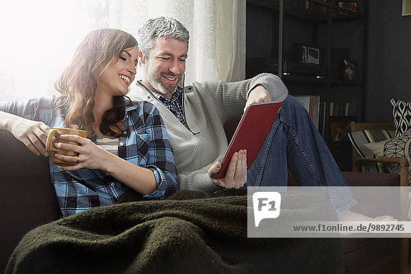 Junges Paar sitzt auf dem Sofa  trinkt Kaffee und schaut sich digitale Tabletten an.