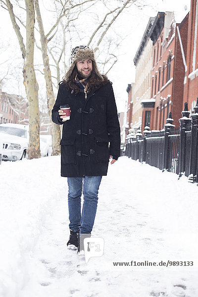 Junger Mann schlendert durch die verschneite Straße und trinkt Kaffee zum Mitnehmen.
