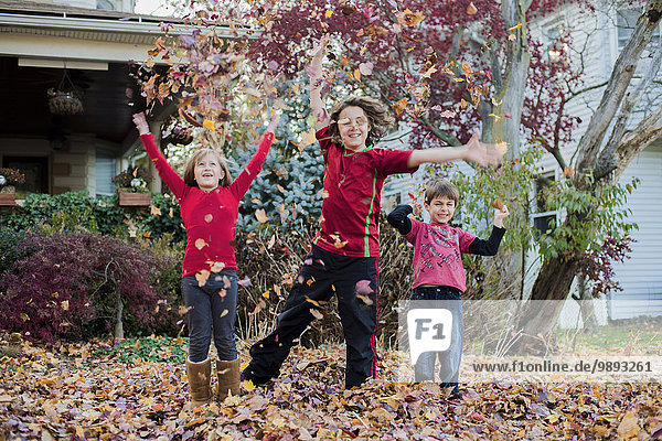 Drei Geschwister spielen mit Herbstblättern im Garten
