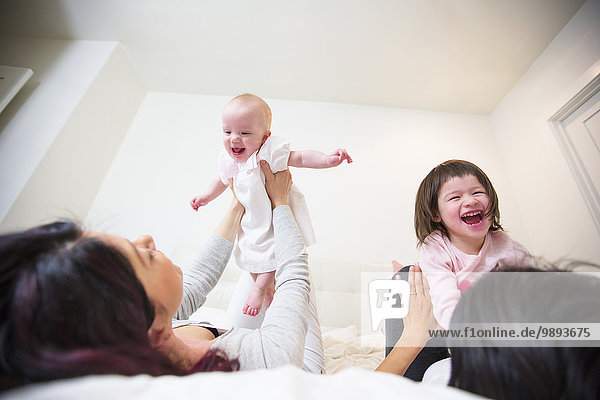 Over the shoulder view of female couple laughing on bed with baby and toddler daughters
