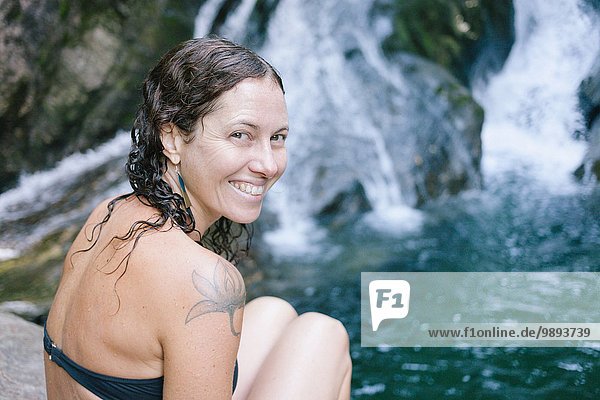 A woman sitting by a swimming hole in woods with waterfall
