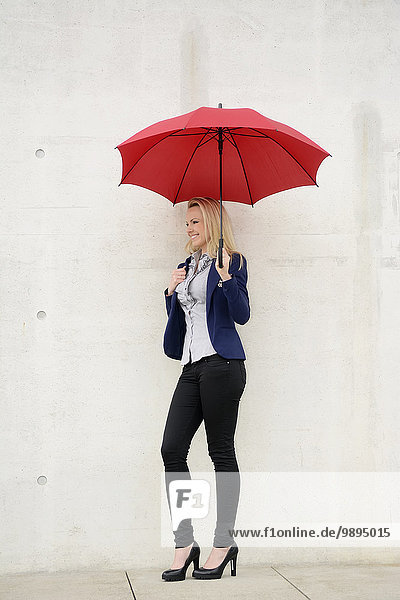Lächelnde Geschäftsfrau mit rotem Regenschirm an Betonwand gelehnt