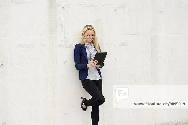 Lächelnde Geschäftsfrau mit digitalem Tablett an Betonwand gelehnt