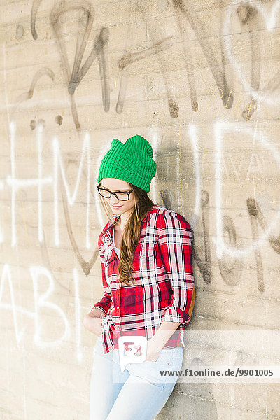 Junge Frau mit kariertem Hemd und grünem Wollhut an der Graffiti-Wand lehnend
