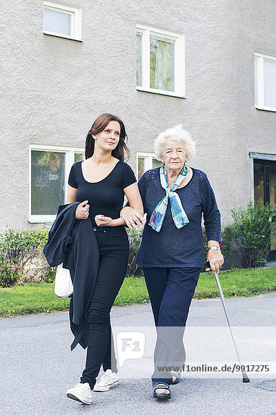 Porträt einer älteren Frau  die den Stock hält  während sie mit ihrer Enkelin auf einem Fußweg gegen den Bau geht.
