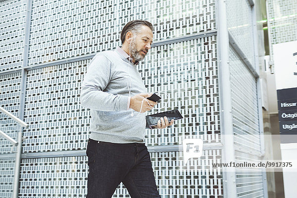 Geschäftsmann mit digitalem Tablett  der Musik über das Handy hört  während er im Büro spazieren geht.