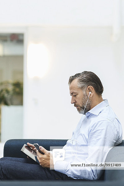 Seitenansicht des Geschäftsmannes mit digitalem Tablett beim Musikhören über Handy in der Bürolobby
