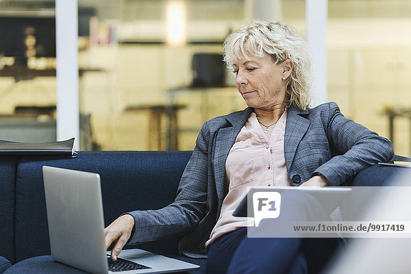 Businesswoman using laptop and digital tablet on sofa at office lobby
