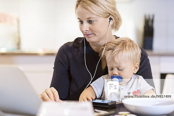 Baby Junge mit Smartphone  während die Mutter Musik auf dem Laptop am Tisch hört.