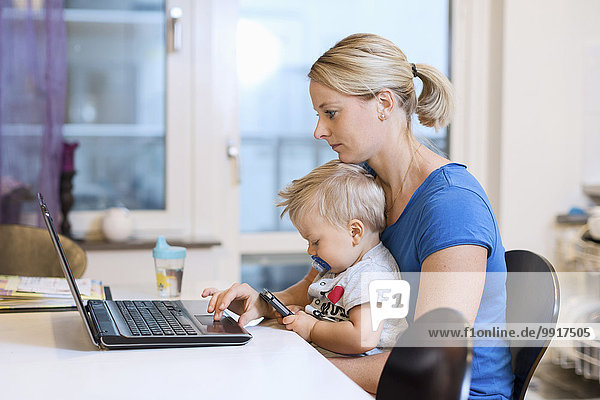 Mutter arbeitet am Laptop  während der kleine Junge zu Hause das Smartphone auf dem Schoß benutzt.