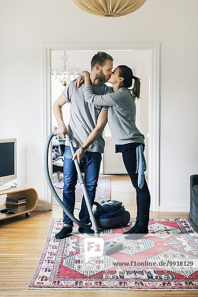 Volle Länge des Mannes  der die Frau küsst  während er den Teppich zu Hause absaugt.