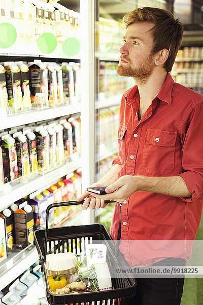 Mann mit Handy beim Einkaufen im Kühlregal im Supermarkt