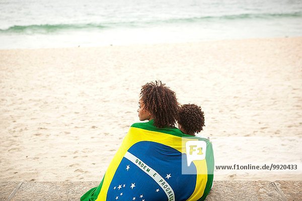 Rückansicht von Mutter und Sohn in brasilianischer Flagge mit Blick vom Strand von Ipanema Rio De Janeiro Brasilien.