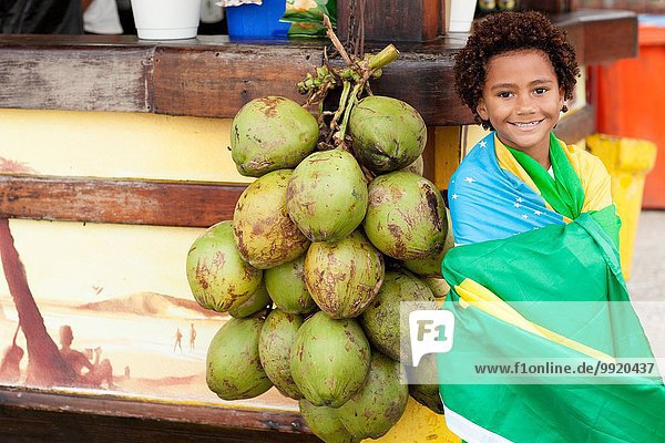 Porträt eines Jungen in brasilianischer Flagge neben einem Haufen Kokosnüsse am Strand von Ipanema  Rio De Janeiro  Brasilien
