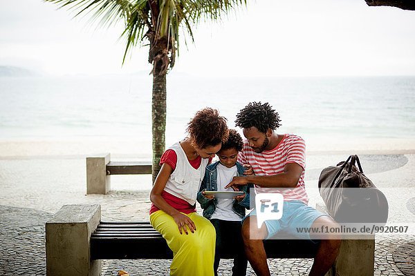Paar und Sohn sitzen auf einer Parkbank mit digitalem Tablett Ipanema Beach Rio De Janeiro Brasilien