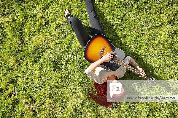 Draufsicht auf eine junge Frau  die auf Gras liegt und Akustikgitarre spielt.