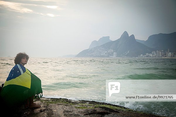 Young man wrapped in Brazilian flag sitting on rock  Ipanema beach  Rio De Janeiro  Brazil