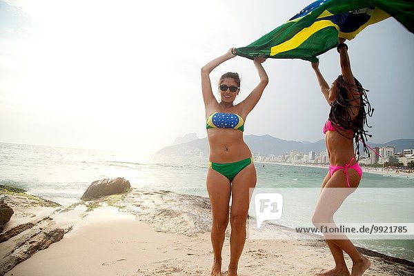Portrait of two young women dancing whilst holding up Brazilian flag Ipanema beach Rio De Janeiro Brazil