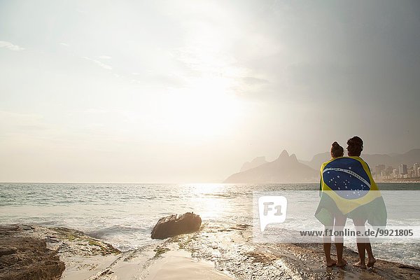 Rear view of two young women wrapped in Brazilian flag on Ipanema beach  Rio De Janeiro  Brazil