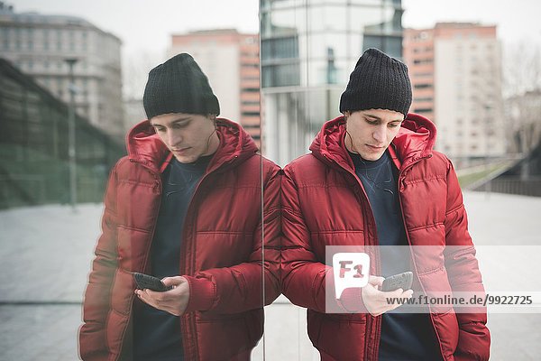 Spiegelbild eines jungen Mannes  der sich gegen ein Bürogebäude lehnt und Smartphone-Texte liest.