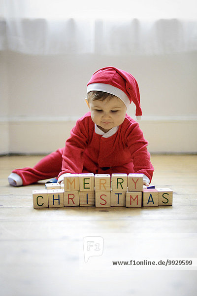 Ein kleiner Junge,  der als Weihnachtsmann verkleidet ist,  mit Spielzeugblöcken,  in denen er Frohe Weihnachten liest.