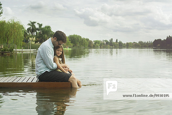 Father and young daughter sitting on dock with legs dangling in lake