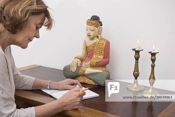 Senior woman writing in notebook  Buddha statue and candles in the background  Munich  Bavaria  Germany