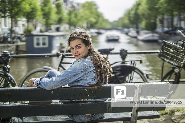 Netherlands  Amsterdam  smiling woman sitting on a bench in front of town canal