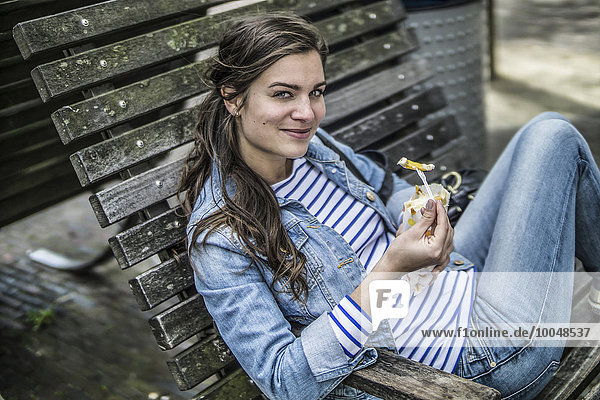 Niederlande  Amsterdam  lächelnde Frau sitzt auf einer Bank und isst Pommes frites.