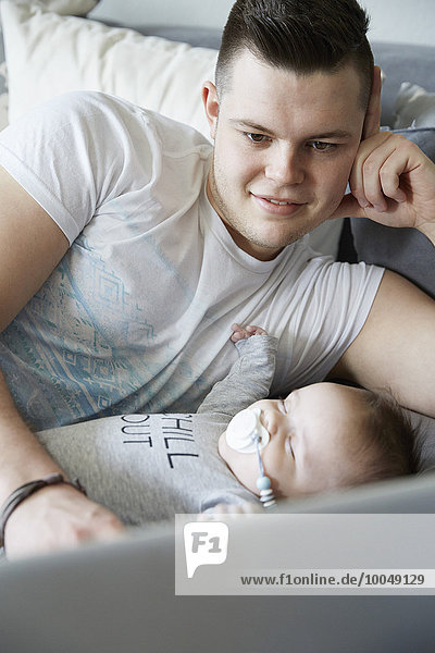 Junger Vater benutzt Laptop mit Baby auf der Couch