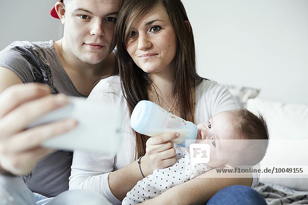 Junger Fatrher nimmt Selfie mit Mutter Flasche fütterndes Baby zu Hause
