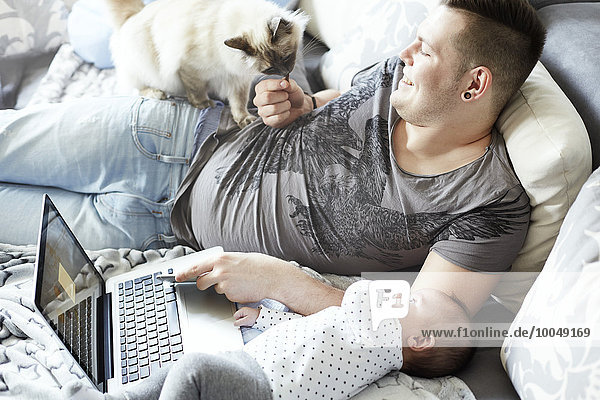 Junger Vater mit Laptop mit Baby und Katze auf der Couch