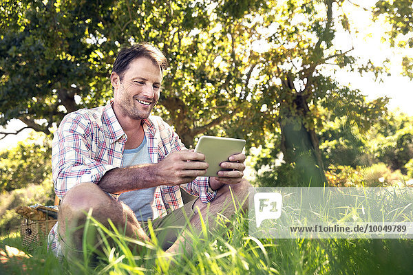 Lächelnder Mann mit digitalem Tablett auf der Wiese sitzend