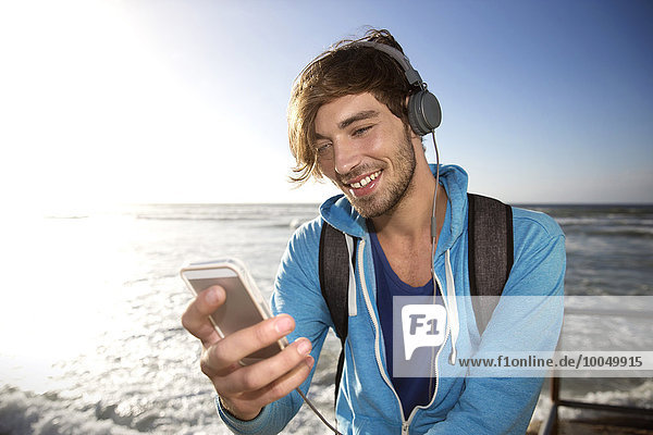 Glücklicher junger Mann an der Küste hört Musik vom Smartphone