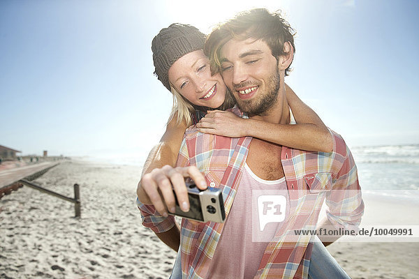 Junge Frau auf dem Rücken ihres Freundes am Strand  die einen Selfie nimmt.