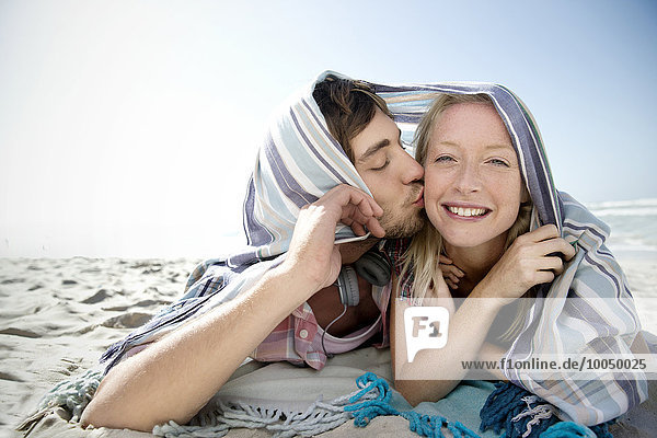 Happy young couple lying on beach kissing under a blanket