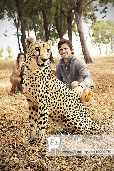 Südafrika  Mann streichelt zahmen Gepard auf der Wiese mit Frau im Hintergrund
